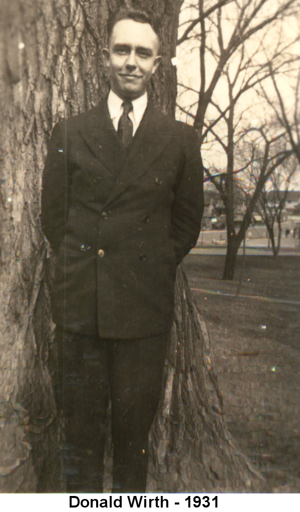 Black and white photo of Donald Wirth, a thin young man with thinning hair in a black suit standing in front of a thick tree trunk.